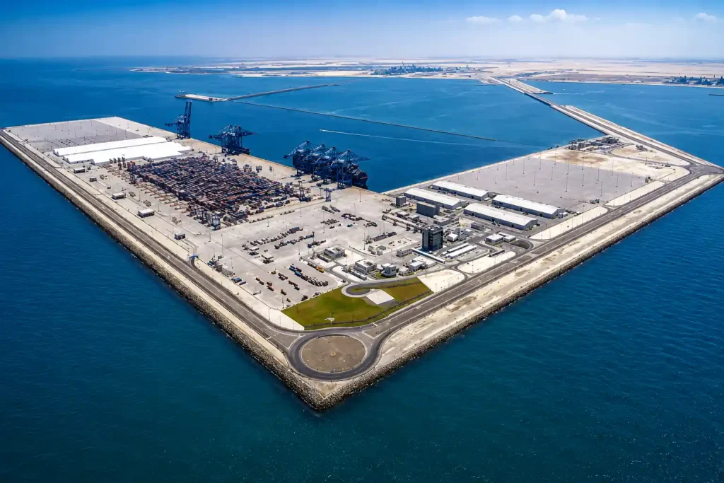 Aerial view of a modern industrial port island with shipping containers, cranes, warehouses, and surrounding blue waters.