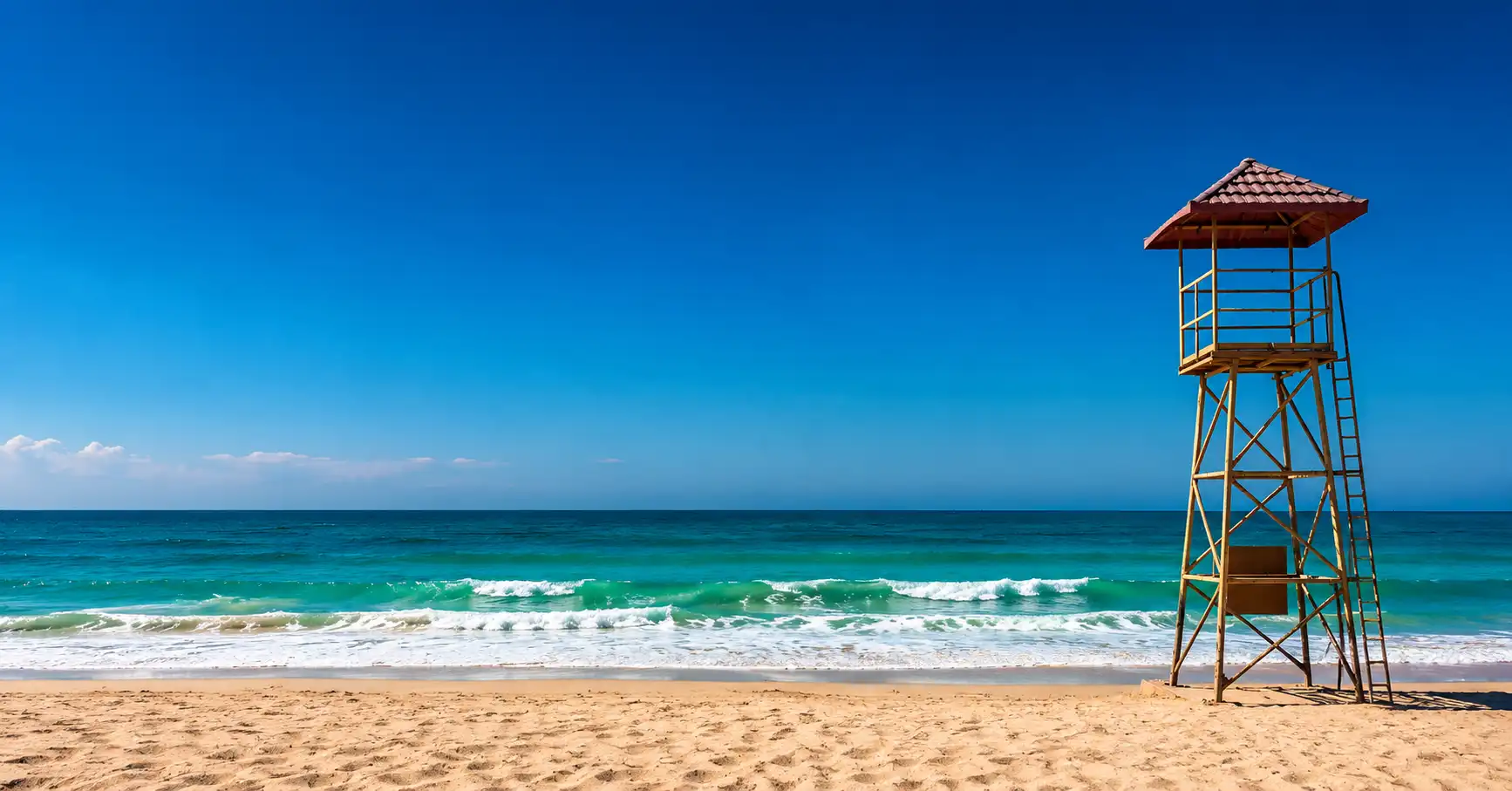 Beautiful panoramic view of Ajman Corniche beach featuring soft sandy shores, turquoise sea waves, clear blue sky, and a lifeguard tower overlooking the peaceful coastline.