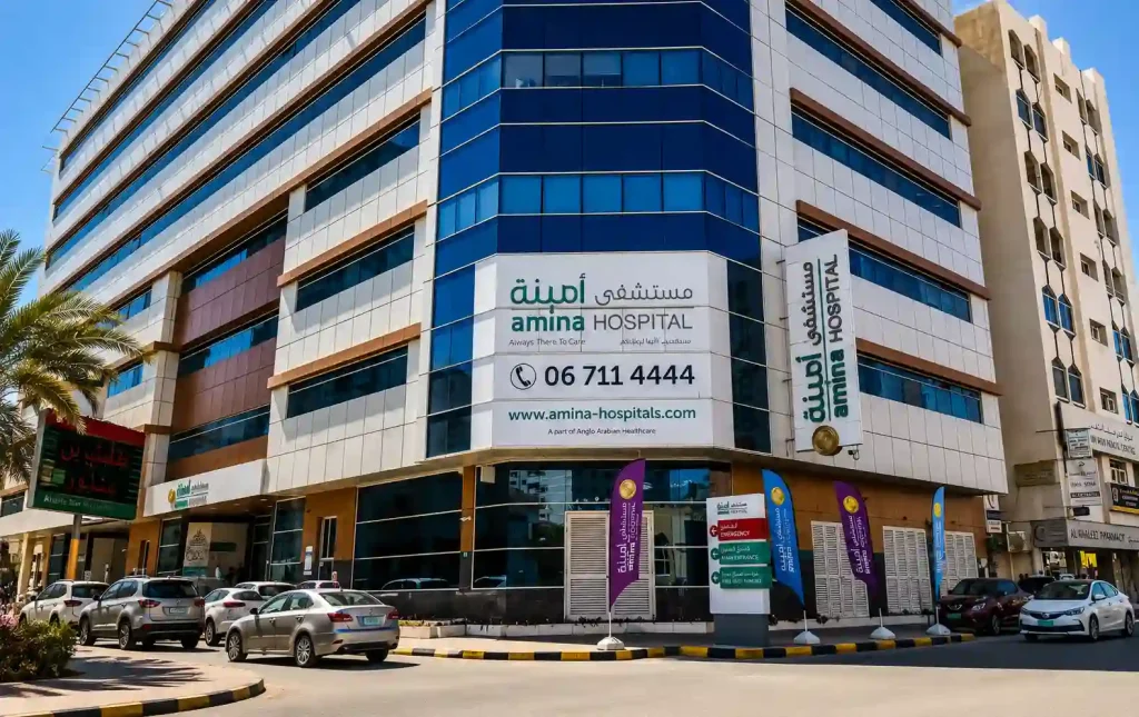 Modern exterior view of Amina Hospital featuring a multi-story glass-front building, emergency entrance signage, parked cars, and a clean urban street scene under a bright blue sky.